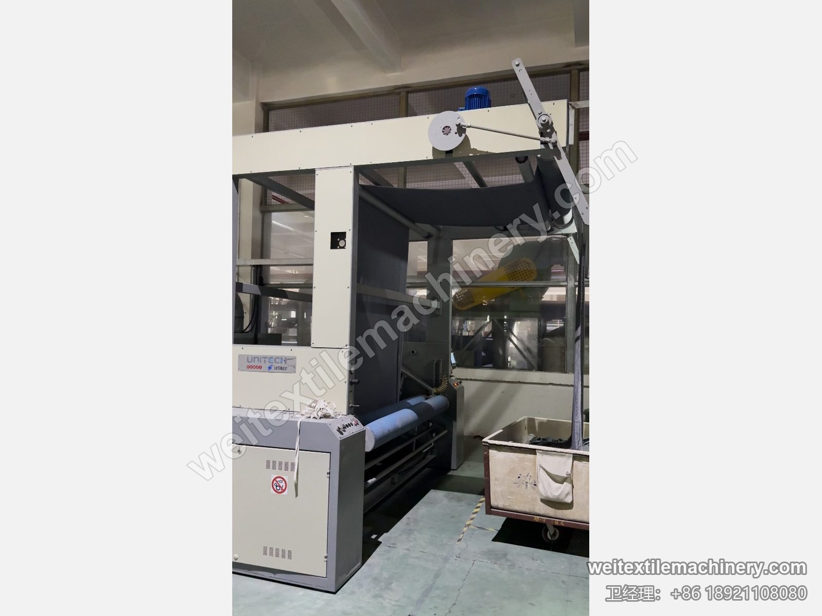 二手 Unitech F2 链条式汽蒸预缩机机端单元，UNITECH Santalucia 标牌、高位导布架挂深色织物与布车